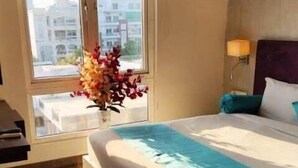 A hotel room with a bed, a window with curtains, a wall-mounted air conditioner, and a vase with flowers. - Hotel Gurveer Royal (Lucknow)