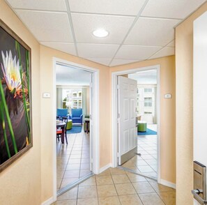 Ein Hotelkorridor mit Fliesenboden, einem Blumengemälde und einer Tür zu einem Zimmer mit blauem Sessel und Tisch.