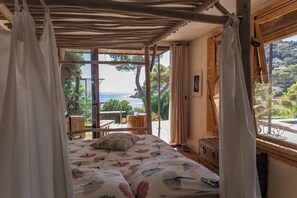 Un dormitorio con cama, cortinas, una ventana con vistas al mar, una mesa de madera y una silla.