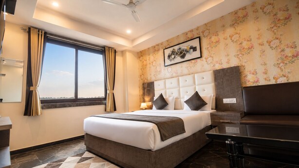 A hotel room with a large bed, a desk, a chair, a window with curtains, a wall-mounted artwork, and a view of the sky. - Hotel Hyfa Continental (Lucknow)