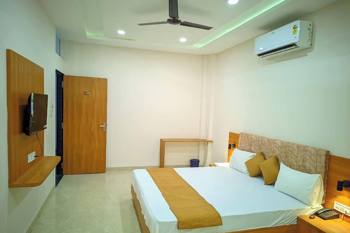 A hotel room with a bed, bedside table, a TV mounted on the wall, an air conditioning unit, and a ceiling fan.