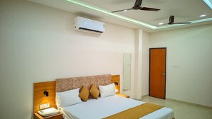 A hotel room with a bed, nightstand, wall-mounted air conditioner, ceiling fan, and a door. - AVR Hotel (Indore)
