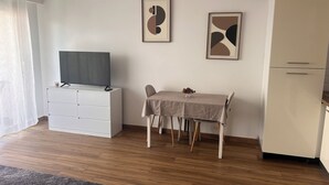 Ein modernes Wohnzimmer mit Fernseher auf einem weißen Schrank, einem Esstisch mit Stühlen und einem Kühlschrank.