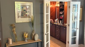 Une chambre avec un plancher en bois, une table de chevet ornée d’un vase et d’un cadre photo, et une porte donnant sur la cuisine.