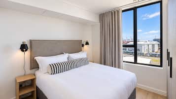 Una habitación de hotel moderna con una cama grande, una mesita de noche, dos lámparas de pared y un ventanal con vistas a edificios industriales.