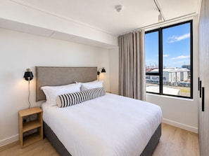 Ein modernes Hotelzimmer mit einem großen Bett, einem Nachttisch, zwei Wandleuchten und einem Fenster mit Blick auf Industriegebäude.