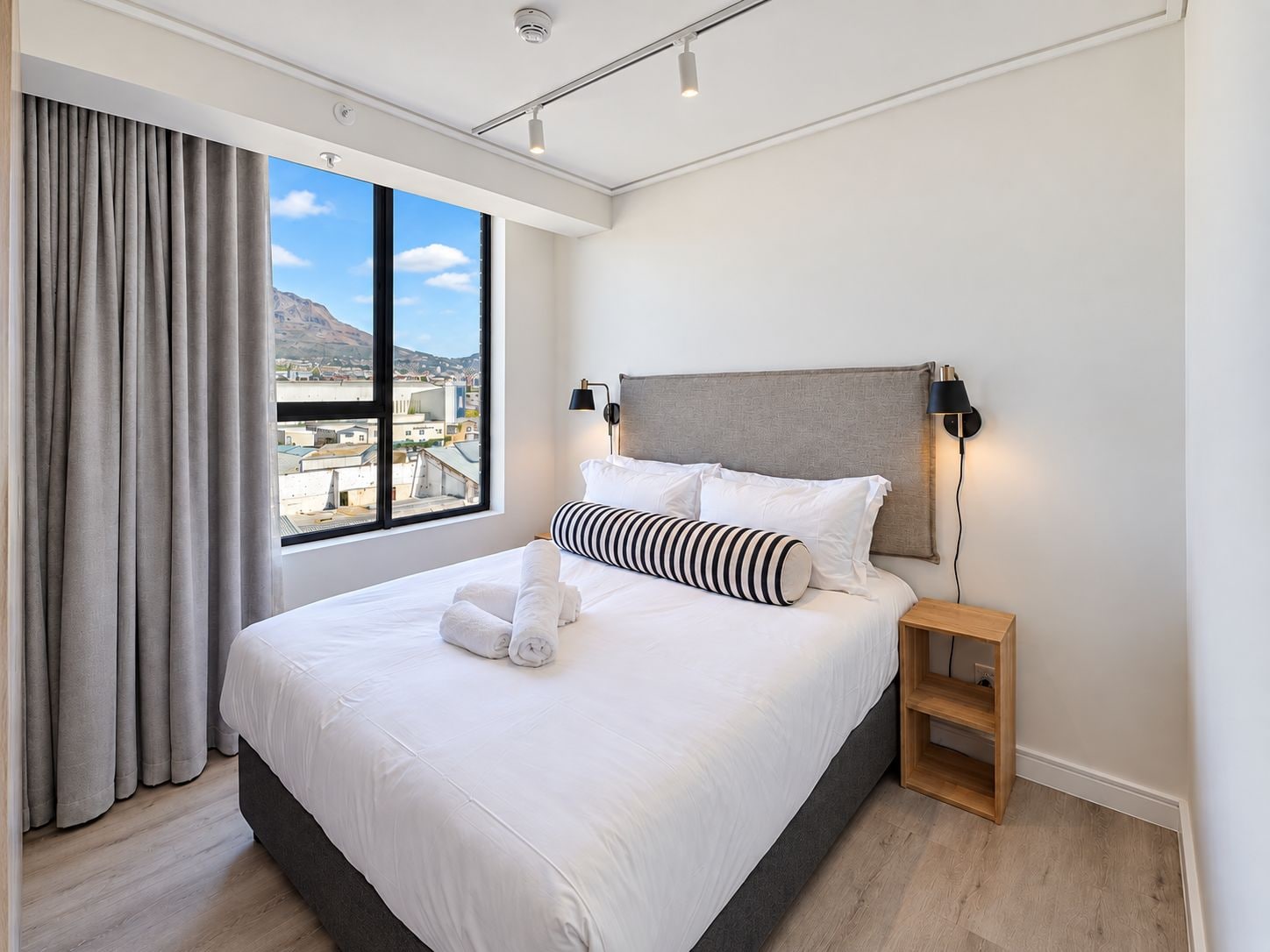 Habitación de hotel moderna con una cama grande, una mesita de noche, una lámpara y vistas a un paisaje urbano a través de un ventanal amplio.