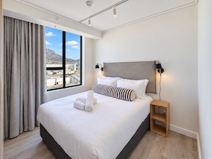 A modern hotel room with a large bed, a nightstand, a lamp, and a view of a cityscape through a large window. - Maritime Place (Cape Town)