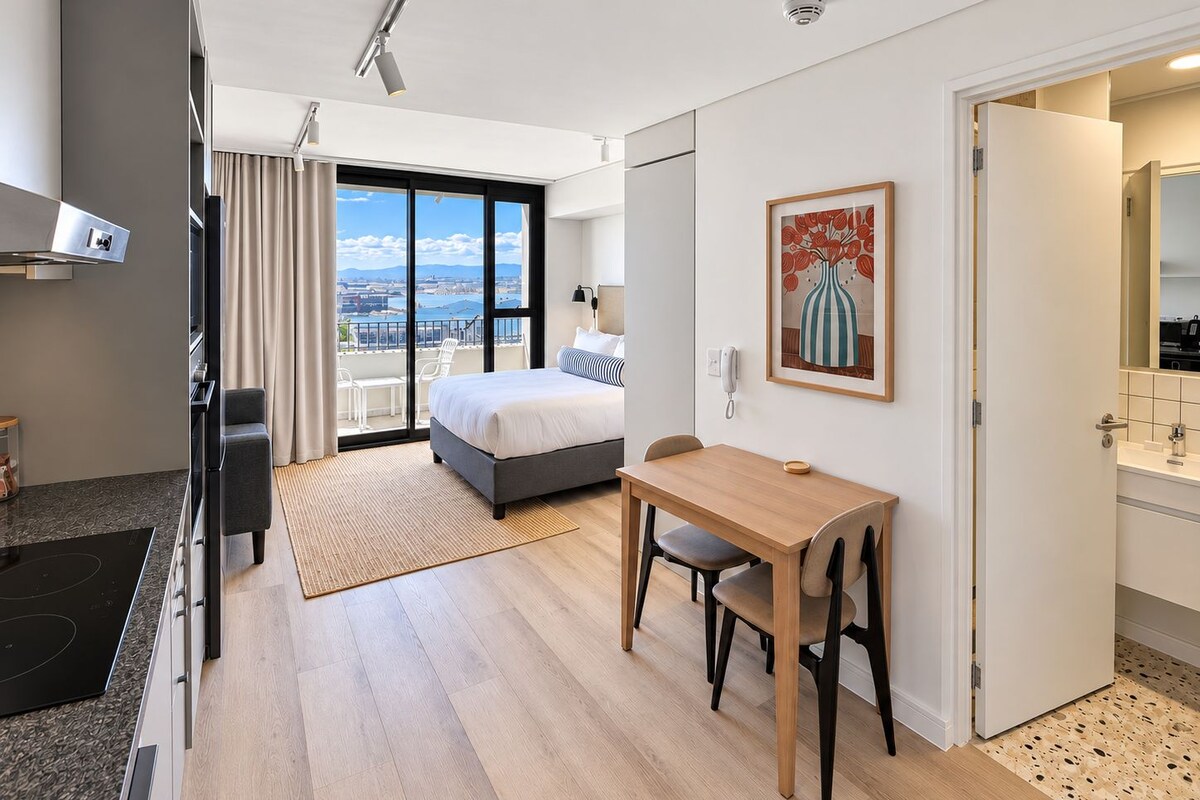 A modern hotel room with a bed, a dining table, a view of the city, and a kitchenette.