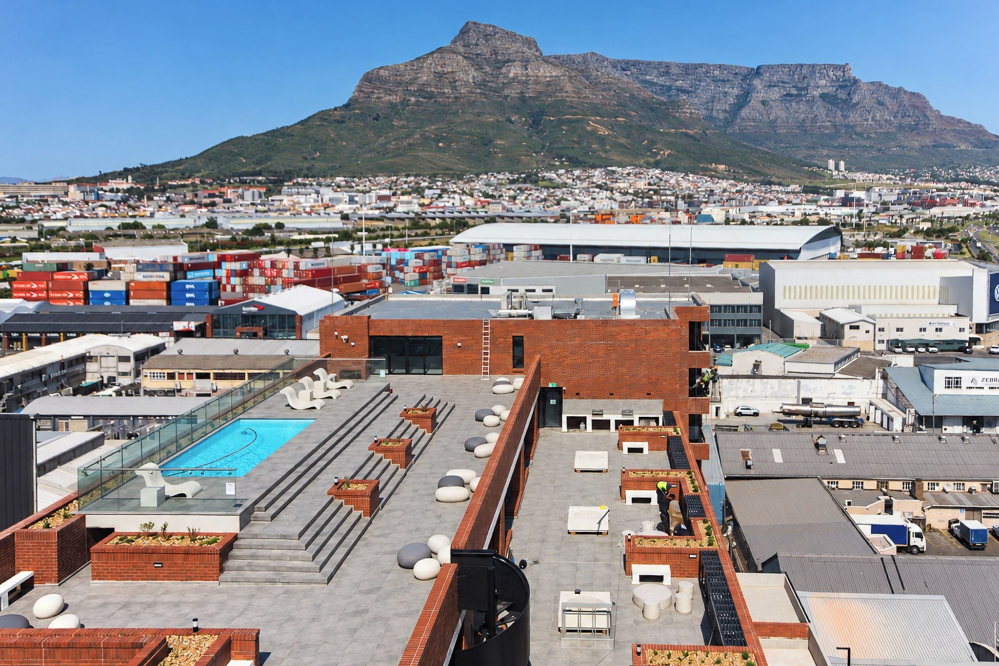 A rooftop with a swimming pool, seating areas, and a view of a cityscape and mountains in the background.