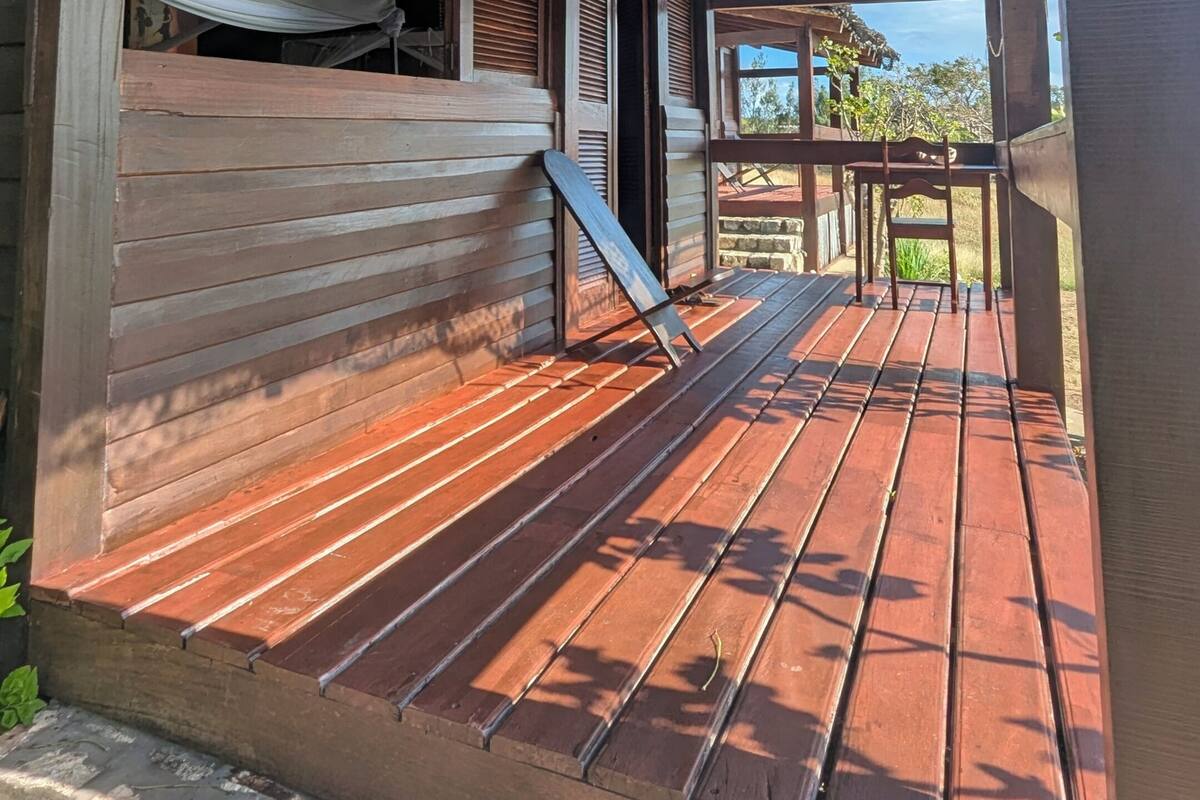 Una terraza de madera con un banco, un área techada y vistas a la vegetación al fondo.