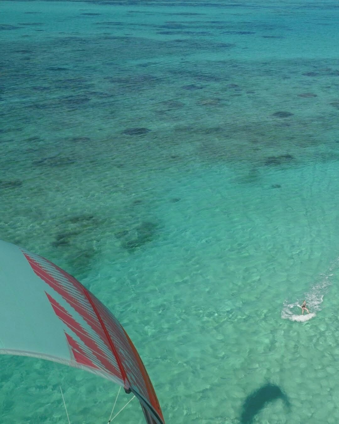Vista despejada de una playa con aguas poco profundas, una persona surfeando y parte de un velero.