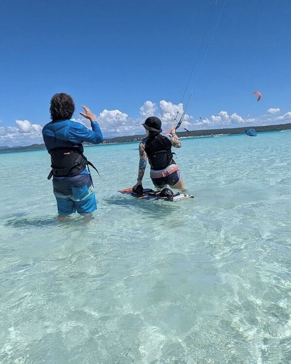 Two people in clear water, one standing and one kneeling, both wearing life jackets, with a clear sky and distant land in the background.