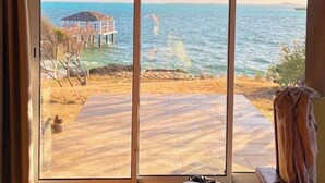 A room with a large sliding glass door leading to a beach view, a thatched roof, and a wooden deck. - Les Paillottes de Babaomby (Antsiranana)