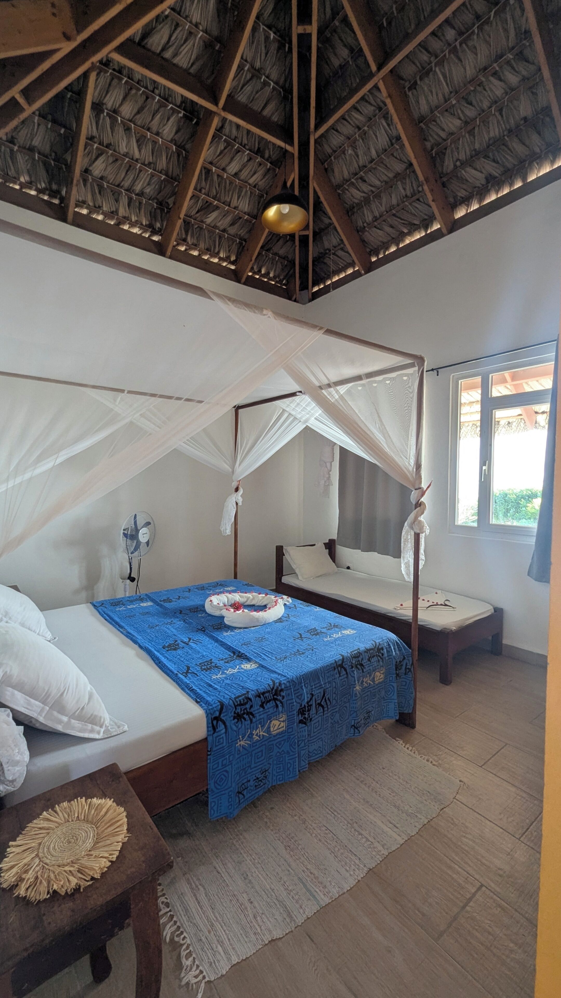 A room with a bed, a bedside table, a standing fan, a window, and a thatched roof ceiling.