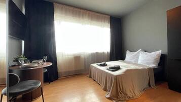 Une chambre d’hôtel avec un lit, une petite table et une chaise, une fenêtre avec des rideaux et un luminaire au plafond.