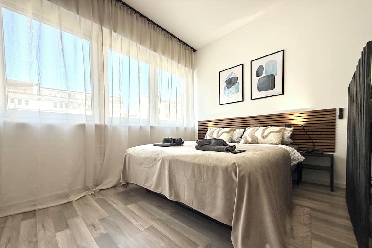 Una habitación de hotel con una cama, una mesita de noche, un ventanal con cortinas y dos cuadros en la pared.