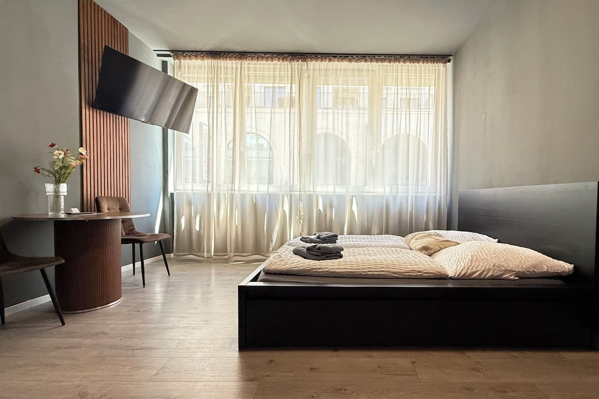 Un dormitorio con una cama, un escritorio, una silla, un televisor y ventanas amplias con cortinas translúcidas.
