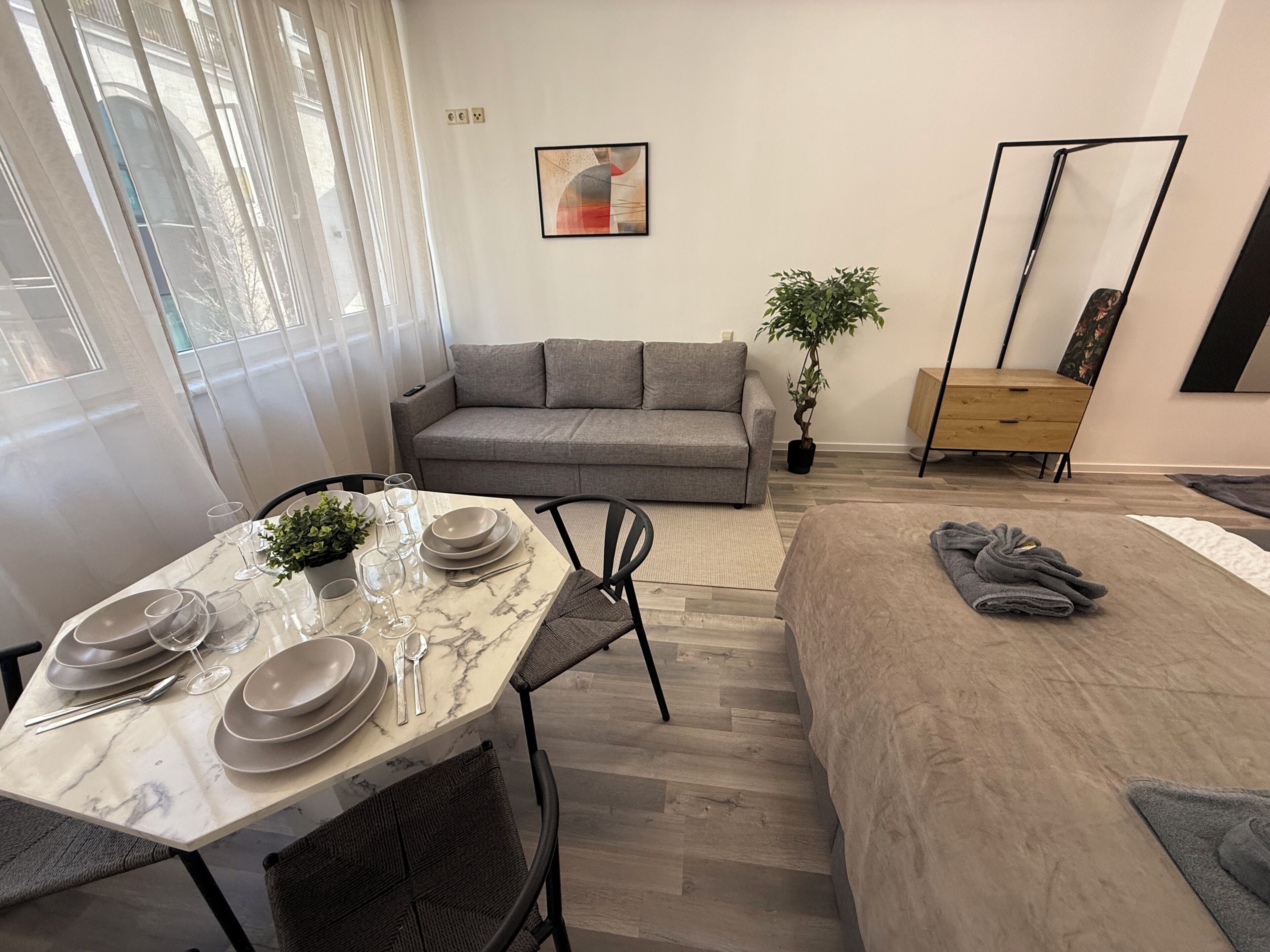 A modern living room with a dining table set for four, a grey sofa, a small potted plant, and a mirror on the wall.
