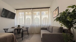 Quarto com sofá, mesa de jantar com cadeiras, televisão fixada na parede e uma janela grande com cortinas translúcidas.