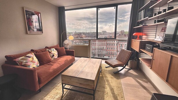 Uma sala de estar moderna com um sofá, uma mesa de centro de madeira, uma poltrona e vista para o horizonte urbano pelas amplas janelas.