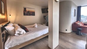 Quarto de hotel com uma cama, uma pintura de paisagem urbana, uma televisão e um espaço de estar com um sofá e uma cadeira.