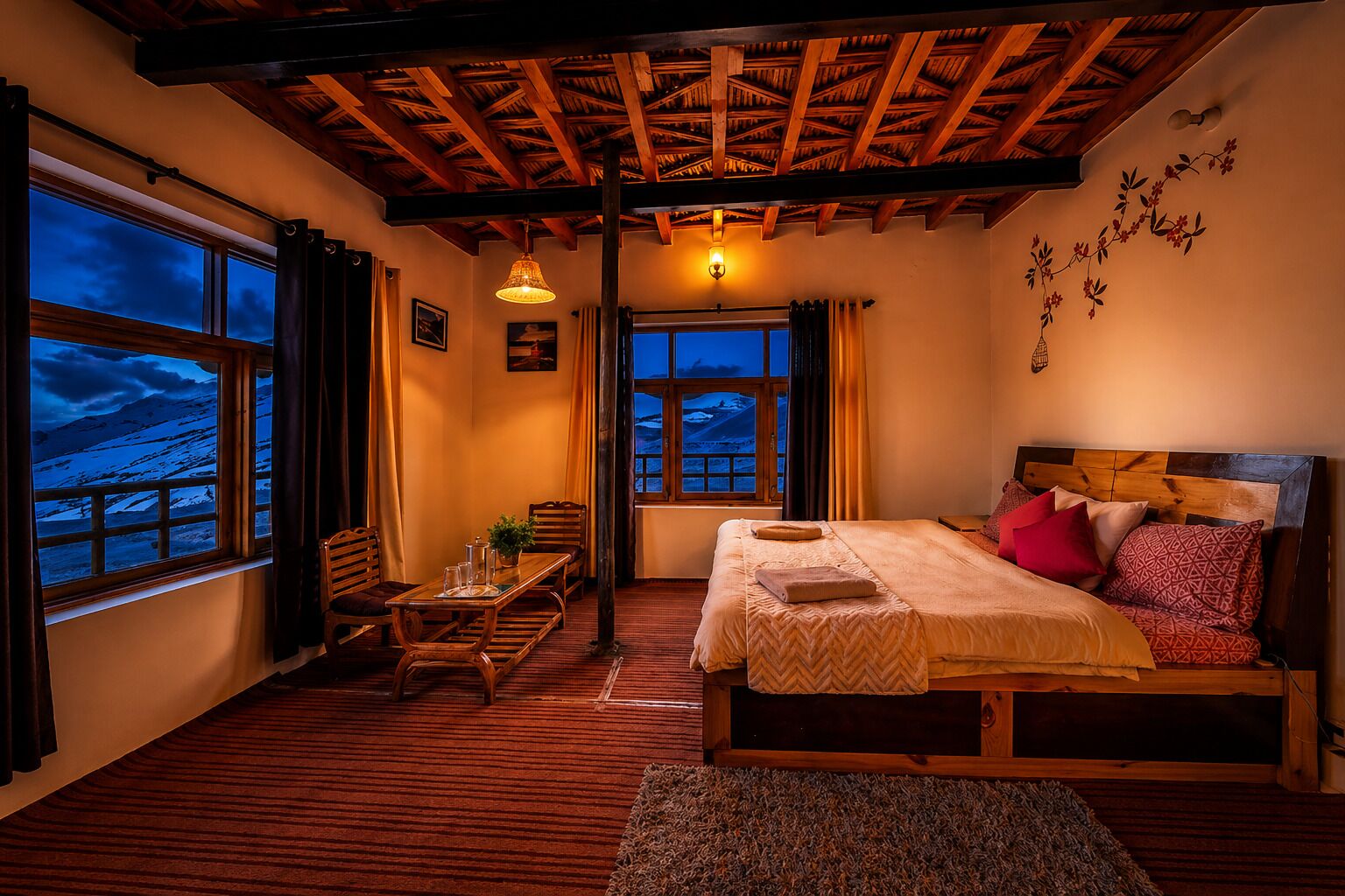A cozy bedroom with a wooden ceiling, a large bed with pillows, a table with chairs, and a view of a snowy landscape through the windows.