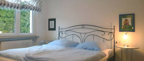 A bed with a metal frame, a bedside table with a lamp, a window with curtains, and a framed picture on the wall.