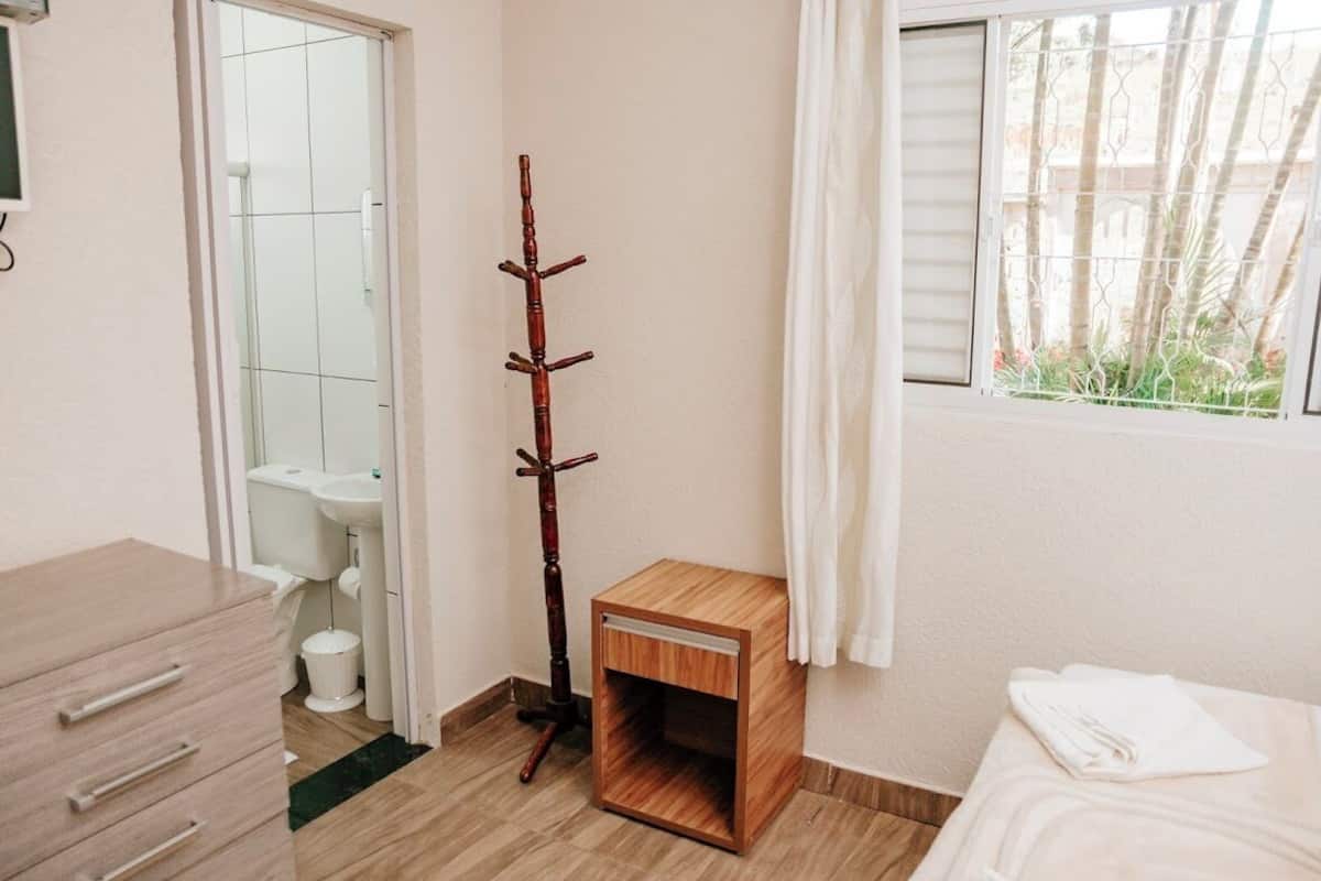 A small, neatly arranged room with a wooden cabinet, a wooden coat rack, a small wooden stool, a bed with a white towel, and a window with a view of greenery.