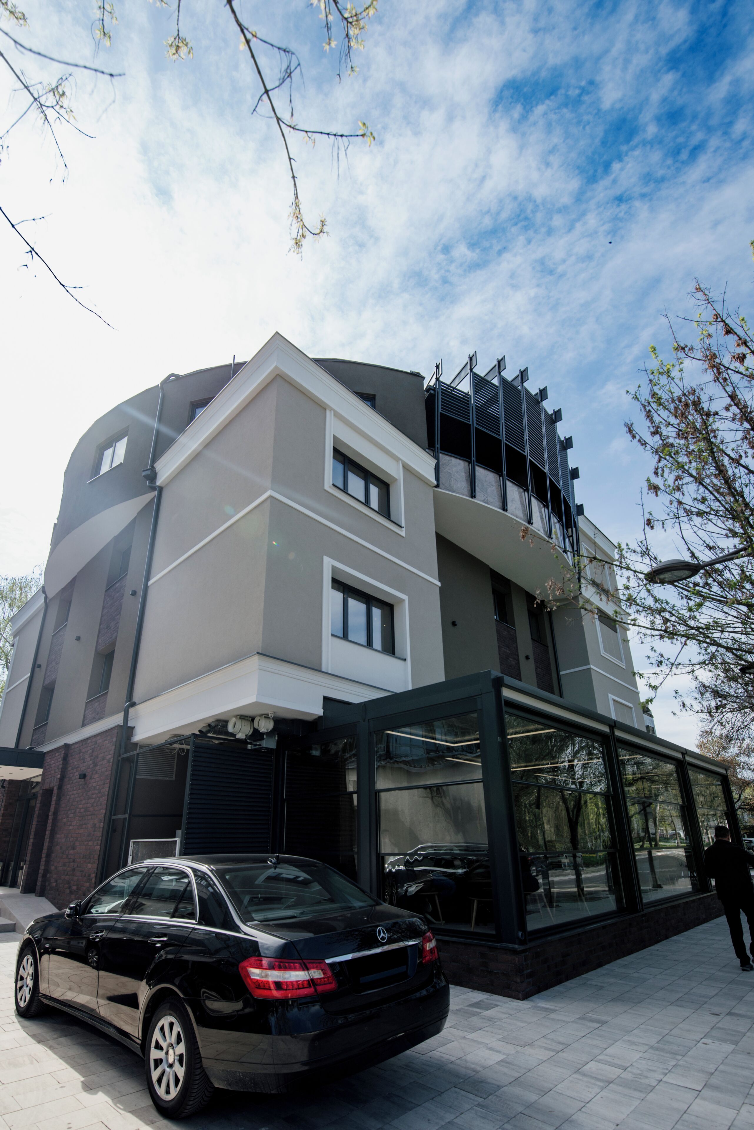 Un edificio moderno con fachada de vidrio, un coche negro estacionado adelante y una persona caminando por la acera.