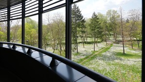 A room with large windows, a curved bench, and a view of a park with trees and a path. - Hotel Botika (Kraljevo)