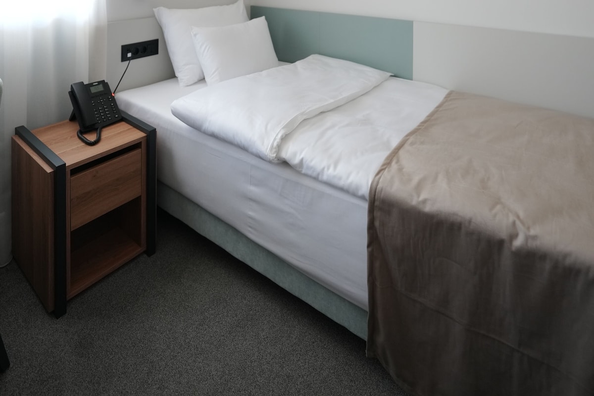 Habitación de hotel con una cama individual, una mesita de noche con teléfono, una lámpara de pared y un cuadro en la pared.