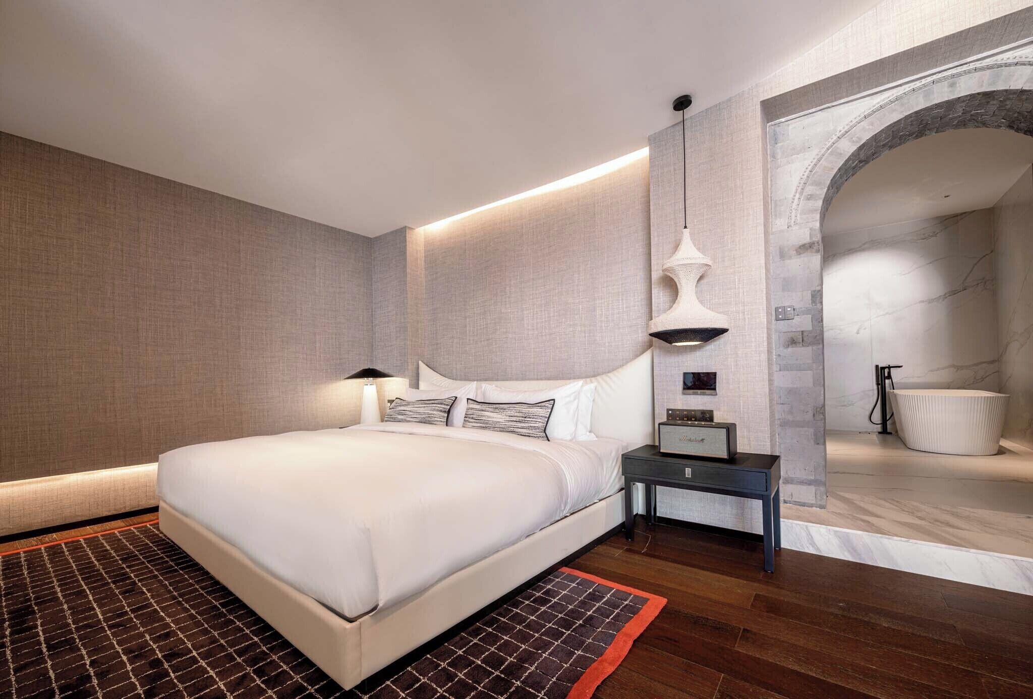 A modern hotel room with a large bed, a nightstand, a wall-mounted lamp, and a view of a bathroom through an arched doorway.