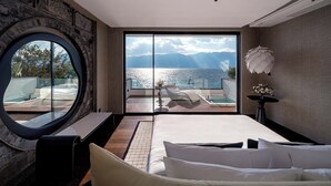 Une chambre avec un grand lit, un miroir rond et une vue sur une étendue d’eau, visible à travers une porte vitrée.