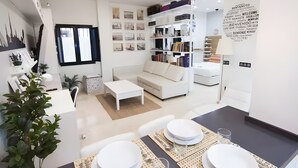 A modern living room with a sofa, coffee table, dining table set for two, and a bookshelf filled with books. - Designer Loft at Seville's Alameda — Smart Tech & Wellness Oasis (Sevilla)