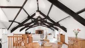 An attic-style room with exposed wooden beams, a round wooden dining table, and a kitchen area with white appliances. - Ivy Cottage, idyllic coastal retreat with sea views (Bideford)