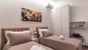 A hotel room with two beds, a wall-mounted air conditioner, a shelf with a basket, and a floral artwork. - Venezia Urban Rooms (Livorno)