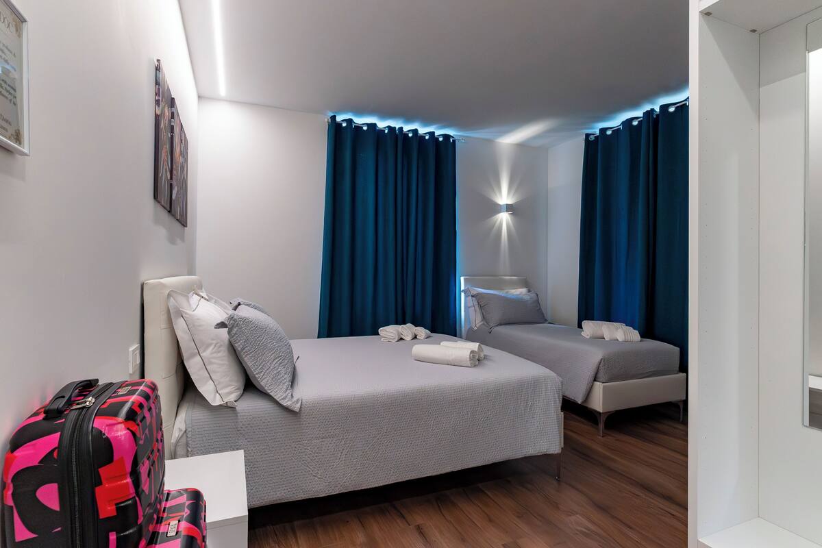 A hotel room with two beds, a desk, a chair, a suitcase, and a wall-mounted light fixture.