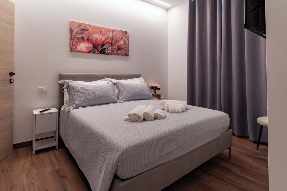 A neatly made bed with pillows, a bedside table with a remote control, and a wall-mounted artwork.