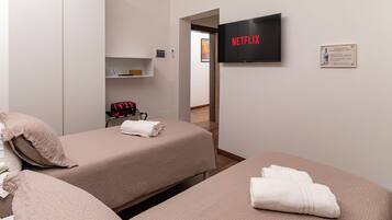 Ein Hotelzimmer mit zwei Betten, einem an der Wand montierten Fernseher, einem Regal mit Gegenständen und einer Tür zu einem anderen Raum.