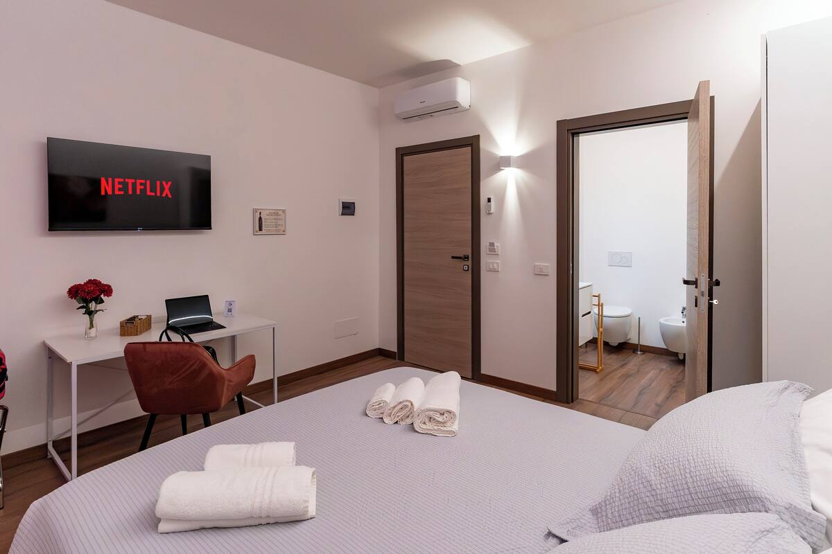 A hotel room with a bed, a desk with a laptop, a TV displaying 'NETFLIX,' a door, and a bathroom visible through an open door.