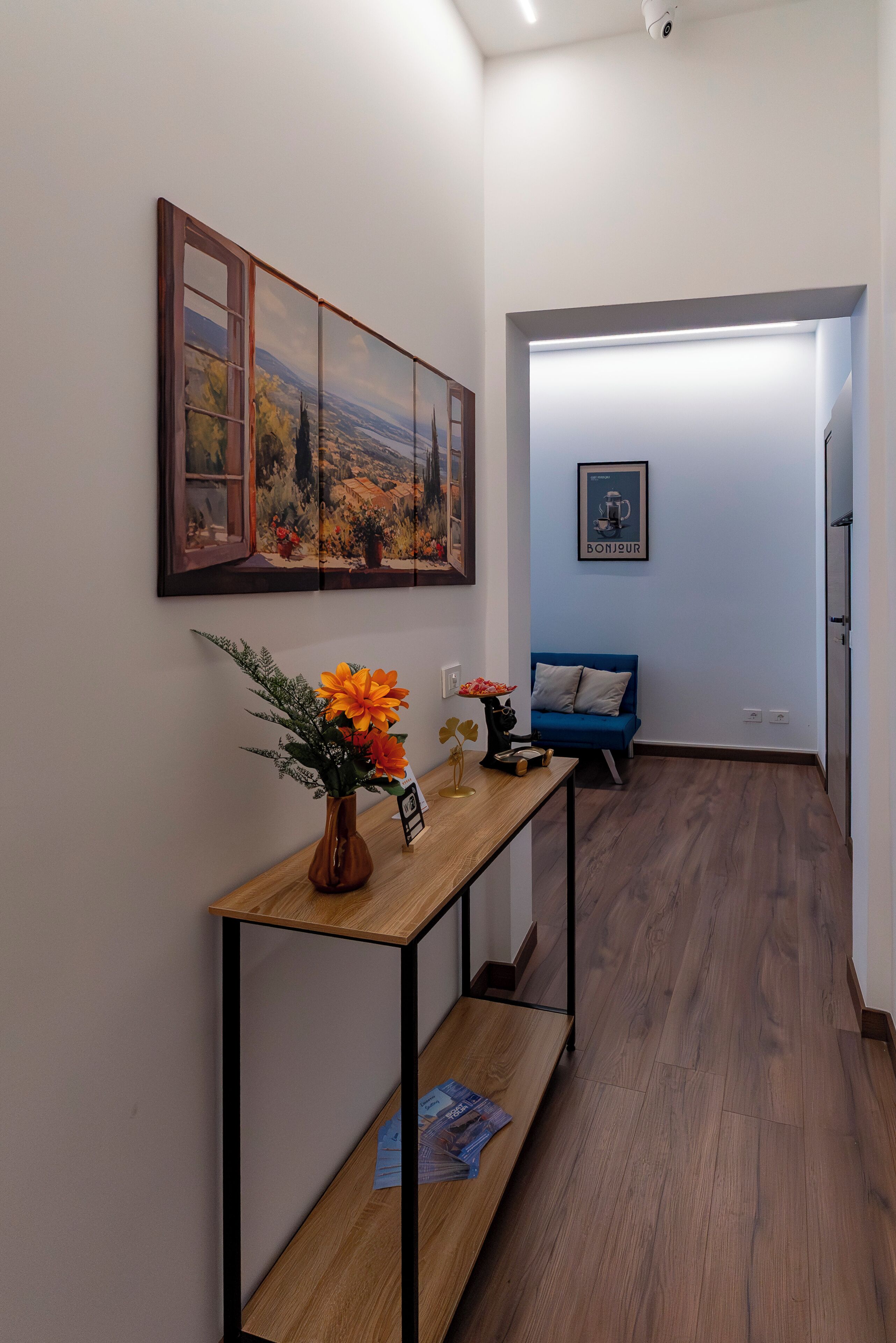 Un corridoio con una panca in legno, un vaso con fiori, un quadro incorniciato e un divano blu.