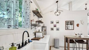 Een moderne keuken met een witte spoelbak, een zwarte kraan en een magnetron. Er staat een houten eettafel met stoelen en een hangende lamp.