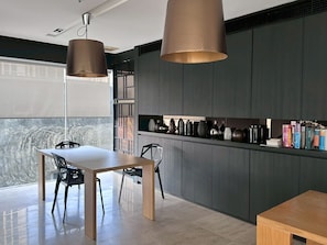 A modern kitchen with dark cabinetry, a dining table, chairs, pendant lights, and a bookshelf with books. - Modern & Cozy Nest 3 Mins to BTS Saphan Khwai (Bangkok)