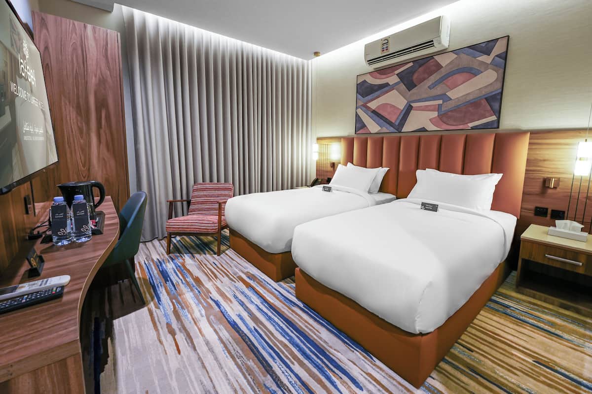 A hotel room with two beds, a desk with a chair, a TV, and a large window with curtains.
