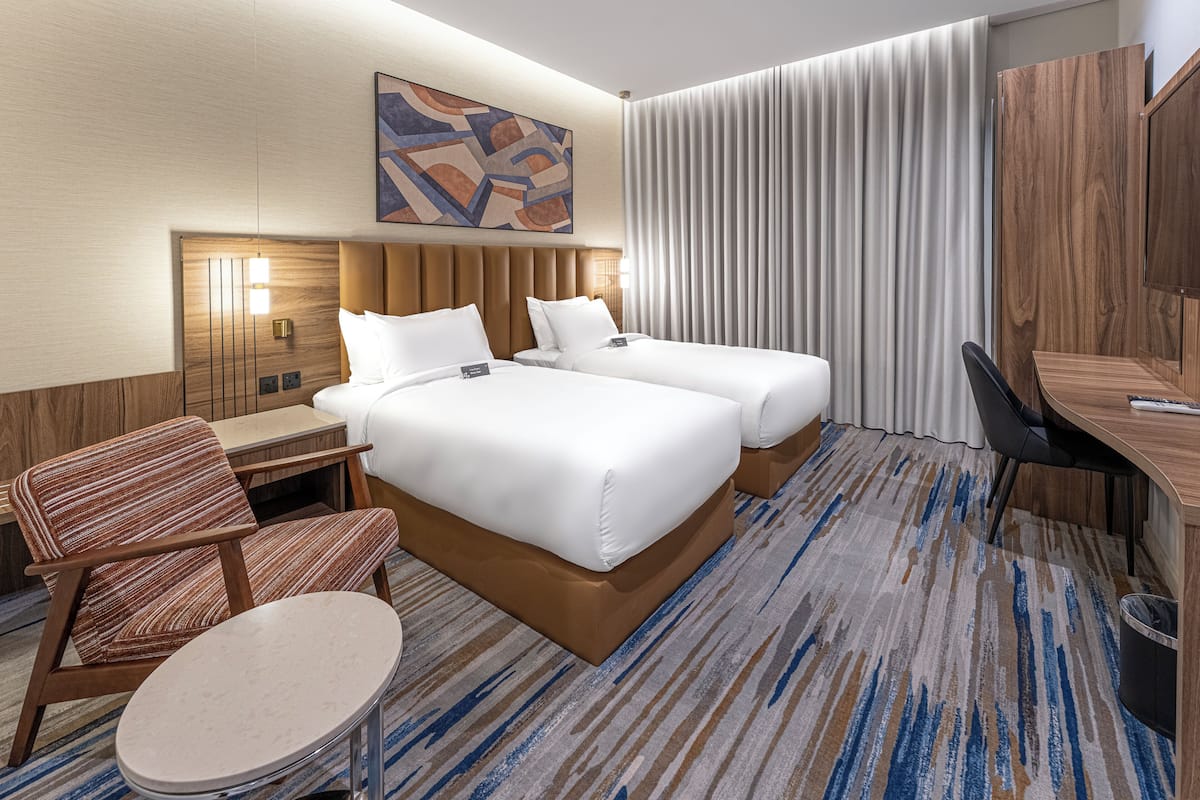 A hotel room with two beds, a desk, a chair, a small table, and a large window with curtains.