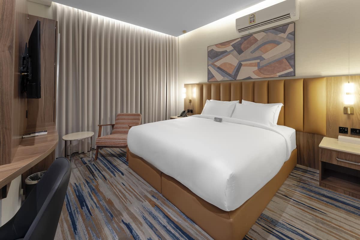 A hotel room with a large bed, a TV mounted on the wall, a desk with a chair, a small table, and a bedside table with a lamp.