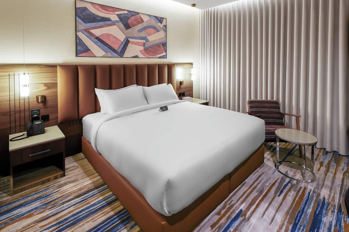 A hotel room with a large bed, a bedside phone, a small table, a chair, a painting on the wall, and a window with curtains.