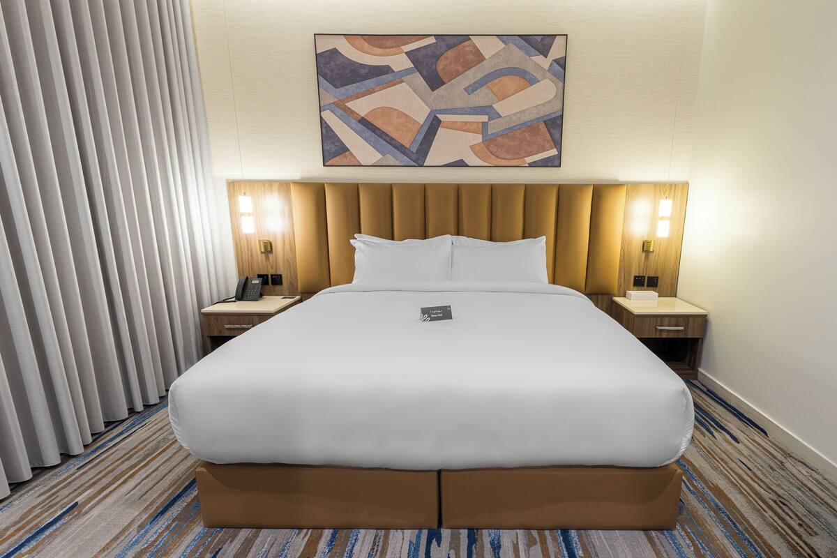 A hotel room with a large bed, a headboard, two bedside tables with lamps, a telephone, and a painting on the wall.
