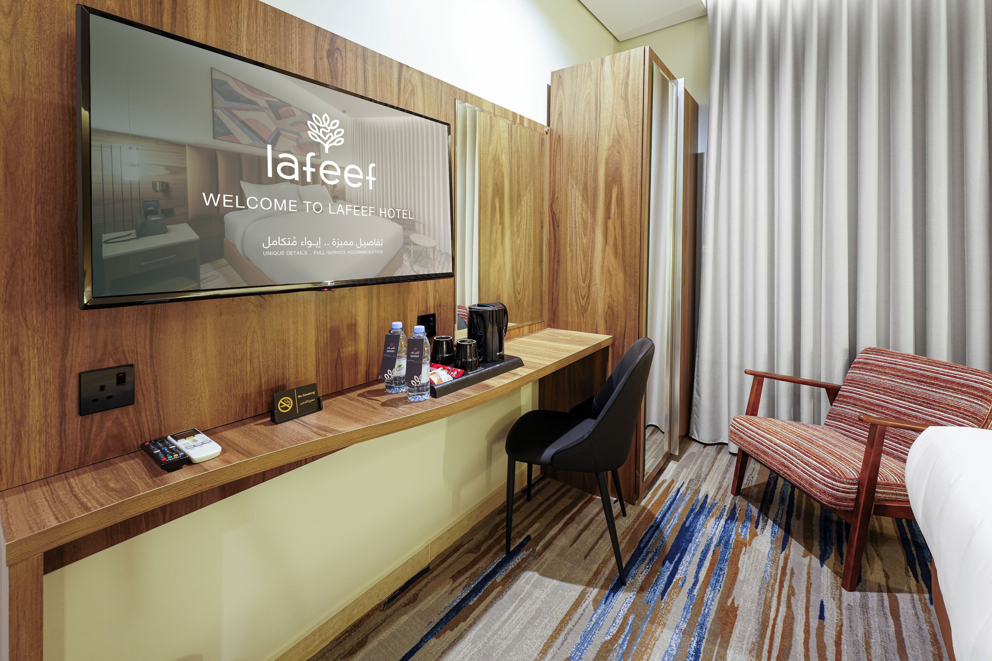 A hotel room with a TV displaying 'LaFeef Welcome to LaFeef Hotel,' a wooden desk with a coffee maker, a chair, and a bed.